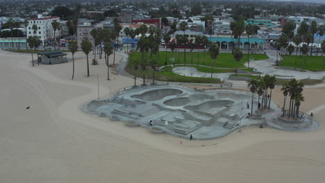 AERIAL:-Turn-around-Venice-Beach-Skatepark-with-Bikers-and-Palm-trees-in-morning,-Cloudy-Los-Angeles,-California